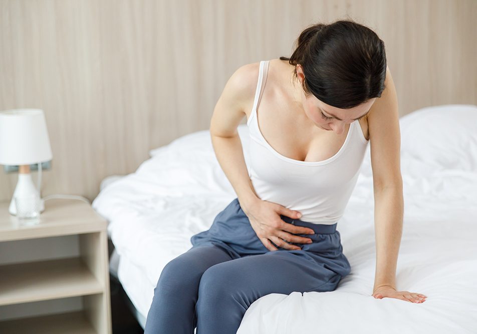 Giovane donna con sintomi del tumore alle ovaie seduta a letto che tocca il basso ventre dolorante con la mano