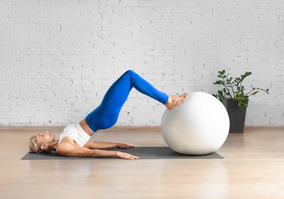 donna-praticando-ginnastica-pelvica-con-fitball Giovane donna caucasica in una spaziosa sala per pilates che pratica ginnastica pelvica con l’ausilio di una fitball