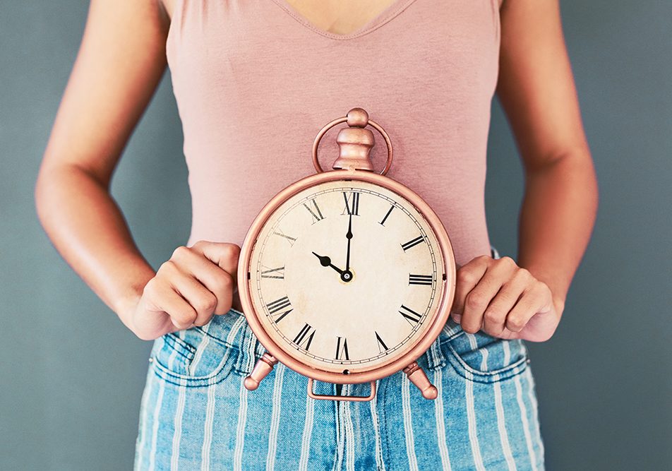 donna-tenendo-sul-grembo-orologio-segnalando-ciclo-in-ritardo-cause Donna con abiti trendy che tiene sul suo grembo un orologio da tavolo per segnalare il problema del ritardo del suo ciclo mestruale e interrogandosi sulle possibili cause