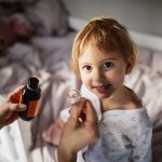 Madre che somministra alla bambina sorridente, adagiata sul letto della sua cameretta, la cefixima prescritta dal pediatra