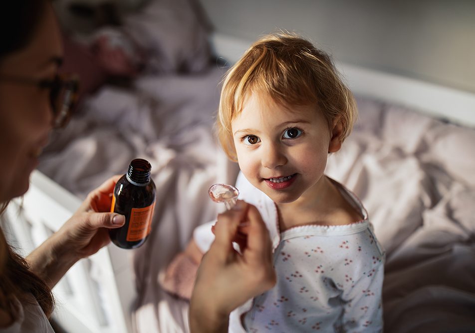 Madre che somministra alla bambina sorridente, adagiata sul letto della sua cameretta, la cefixima prescritta dal pediatra