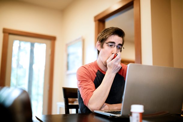 Uomo a casa davanti al computer che utilizza un inalatore per poter gestire i sintomi della fibrosi cistica