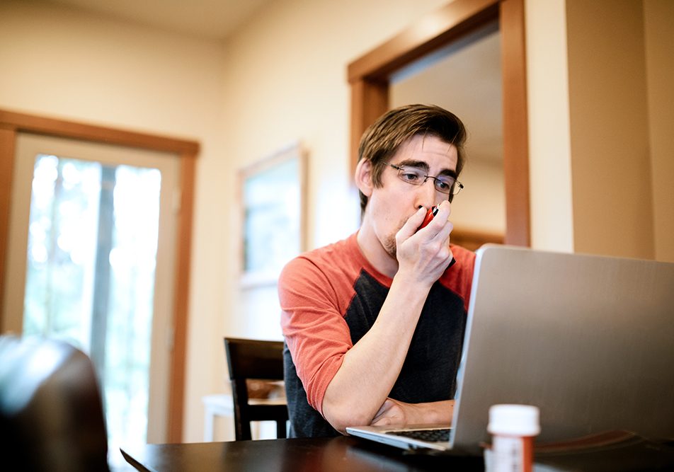 uomo-al-computer-utilizzando-inalatore-per-gestire-fibrosi-cistica-sintomi Uomo a casa davanti al computer che utilizza un inalatore per poter gestire i sintomi della fibrosi cistica