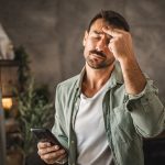 Uomo nel salotto di casa che tiene in mano il suo smartphone e manifesta sintomi da stress