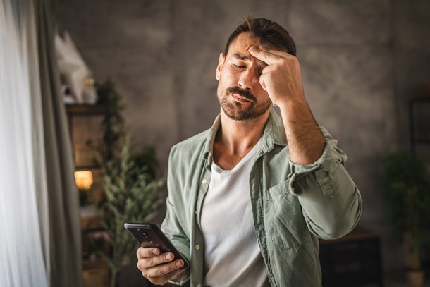 Uomo nel salotto di casa che tiene in mano il suo smartphone e manifesta sintomi da stress