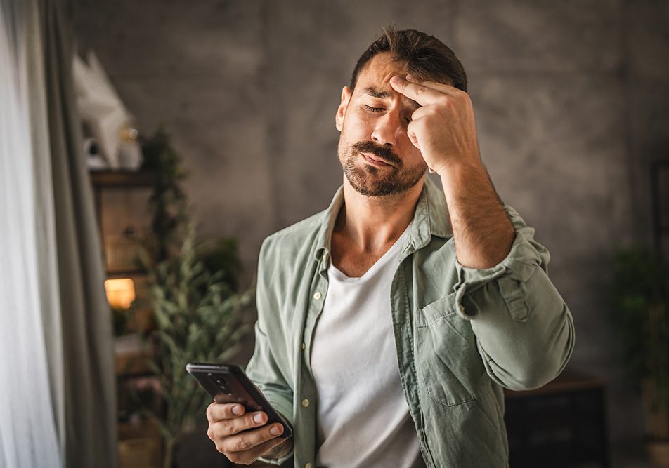 uomo-allo-smartphone-manifestando-stress-sintomi Uomo nel salotto di casa che tiene in mano il suo smartphone e manifesta sintomi da stress