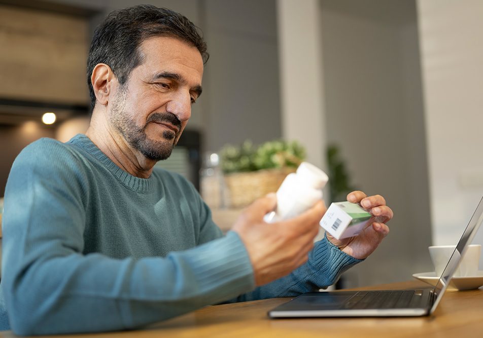 Uomo seduto davanti al computer portatile che legge le confezioni di ketoprofene e cortisone per verificare se è corretto assumerle insieme