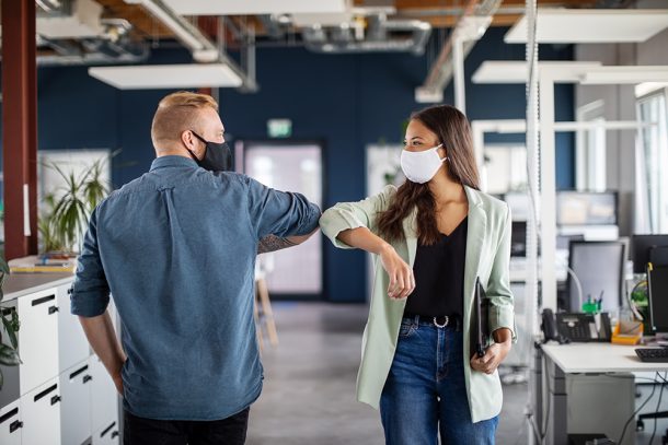 Due colleghi nel corridoio dell’ufficio che si salutano con il gomito, adottando misure di prevenzione per evitare il contagio da Covid-19