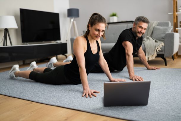 coppia-svolgendo-a-casa-esercizi-per-la-schiena-seguendo-lezioni-online Coppia che svolge degli esercizi per la schiena nel salotto di casa, seguendo delle lezioni online