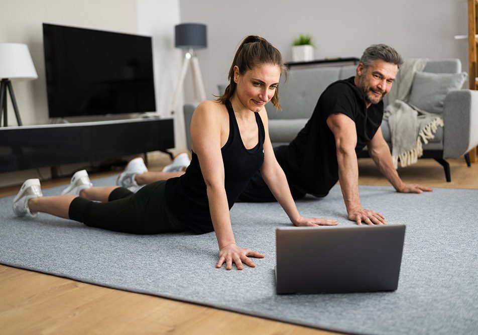 coppia-svolgendo-a-casa-esercizi-per-la-schiena-seguendo-lezioni-online Coppia che svolge degli esercizi per la schiena nel salotto di casa, seguendo delle lezioni online