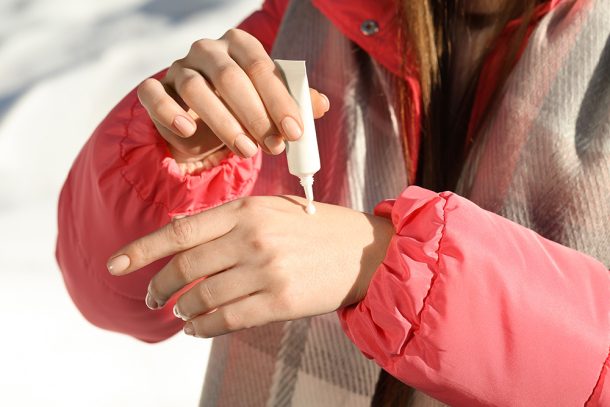 donna-applicando-crema-idratante-per-prevenzione-mani-screpolate Donna che passeggia su un prato innevato, in inverno, mentre applica una crema idratante per la prevenzione delle mani screpolate