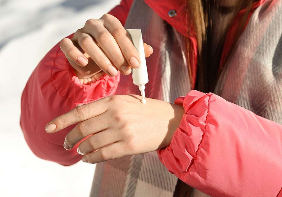 donna-applicando-crema-idratante-per-prevenzione-mani-screpolate Donna che passeggia su un prato innevato, in inverno, mentre applica una crema idratante per la prevenzione delle mani screpolate
