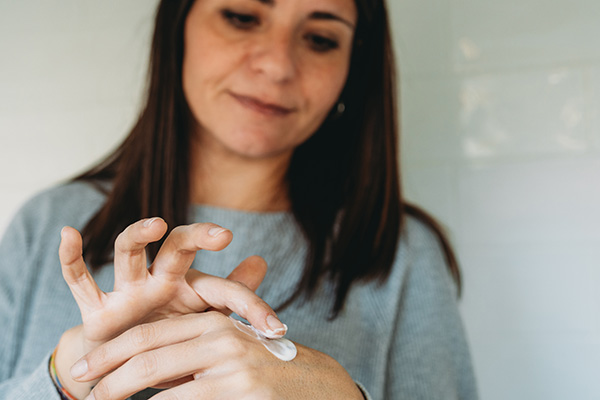 Donna negli interni domestici che soffre di dermatite da contatto mentre applica una crema idratante sulle zone interessate