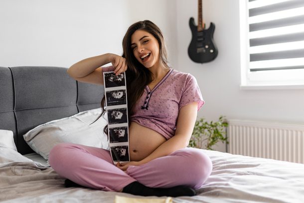 donna-incinta-mostrando-ecografia-dopo-sintomi-gravidanza-primi-giorni Donna incinta in pigiama che mostra felicemente le prime ecografie del nascituro, dopo avere superato i sintomi della gravidanza dei primi giorni