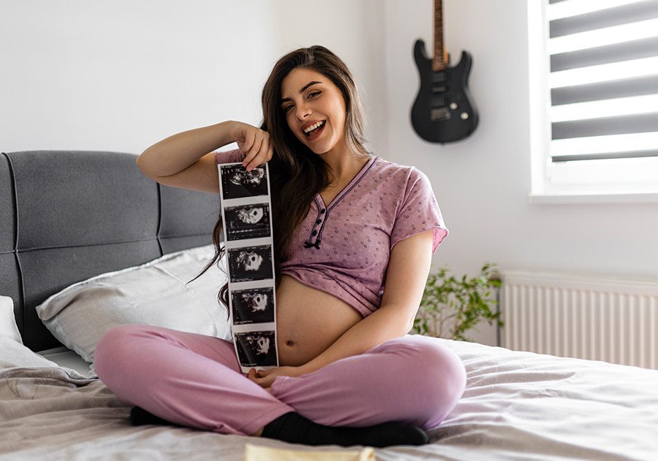 donna-incinta-mostrando-ecografia-dopo-sintomi-gravidanza-primi-giorni Donna incinta in pigiama che mostra felicemente le prime ecografie del nascituro, dopo avere superato i sintomi della gravidanza dei primi giorni