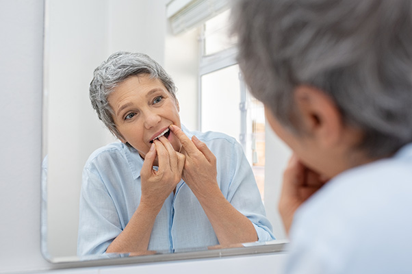 Donna matura nel bagno di casa che osserva la sua dentatura allo specchio mentre segue i suggerimenti del dentista su come pulire la protesi