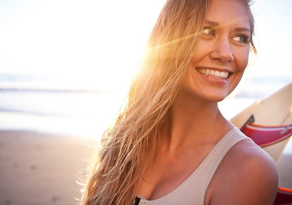 Ragazza dalla chioma bionda che surfa in spiaggia al tramonto, dopo avere applicato una protezione dei capelli dal sole
