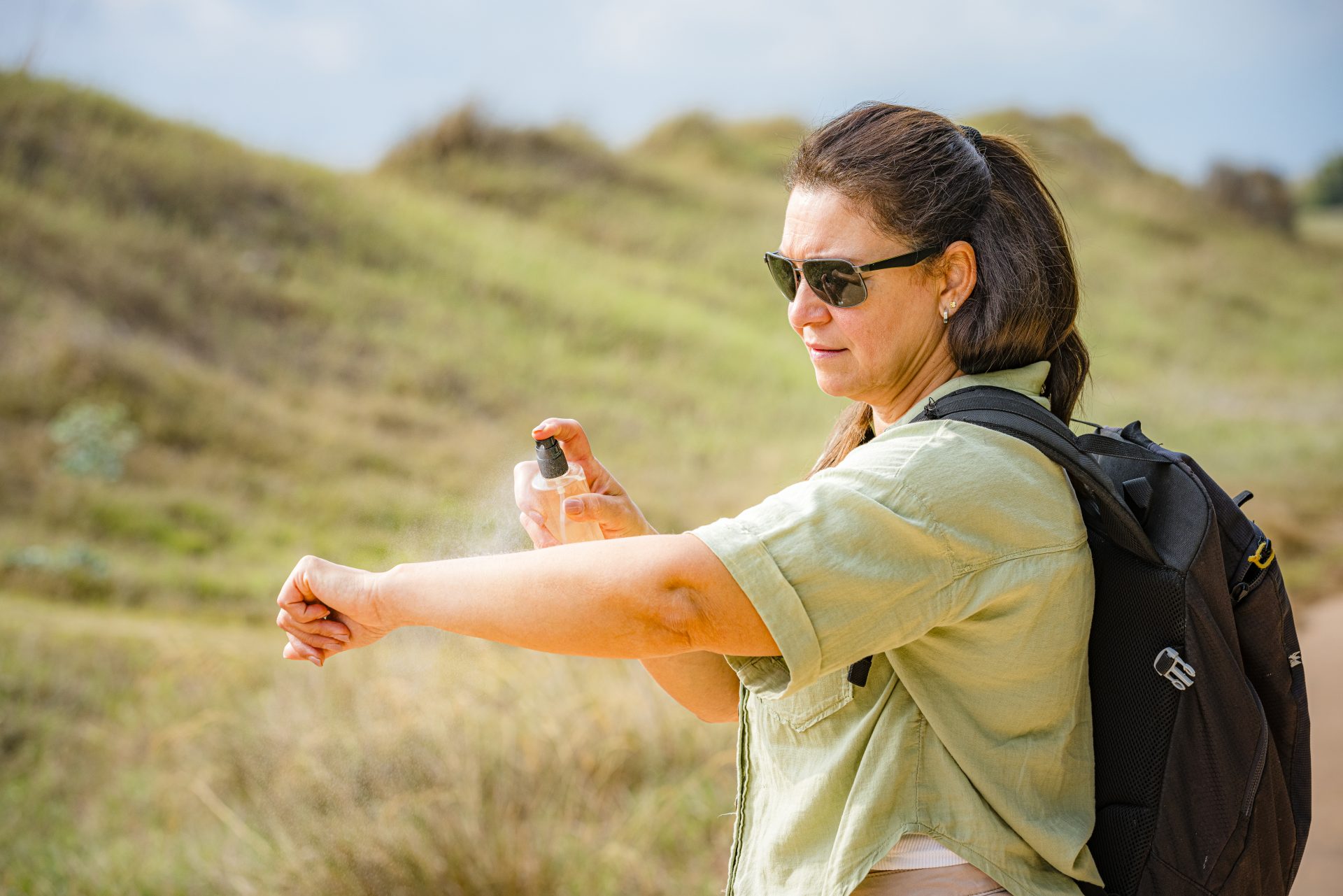 donna con occhiali da sole e zaino spruzza repellente per zanzare sul braccio
