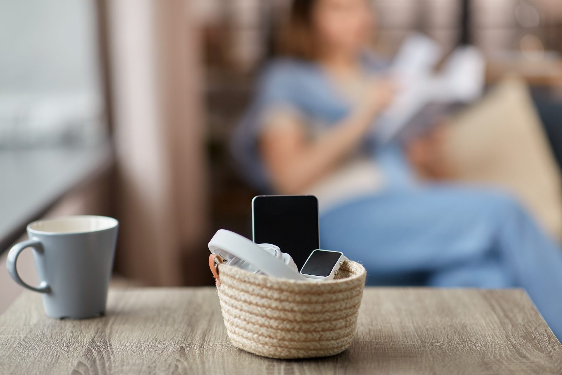 primo piano di tazza e dispositivi digitali dentro a un contenitore con donna che legge sullo sfondo